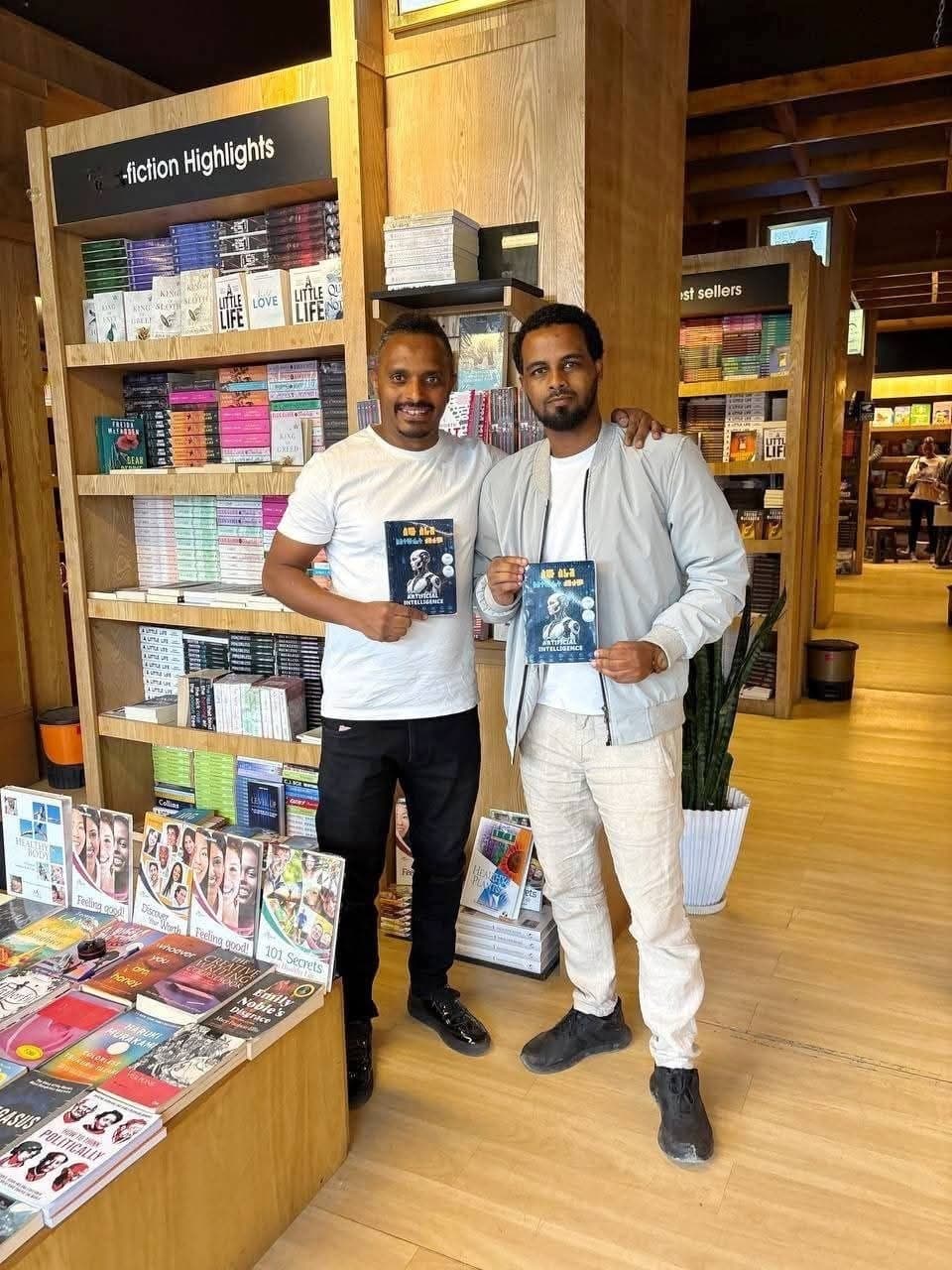 The author holding a printed copy of አርቴፊሺያል ኢንተሊጀንስ ለሁሉም — AI FOR EVERYONE alongside the owner of Jafar Book Center, inside the bookstore