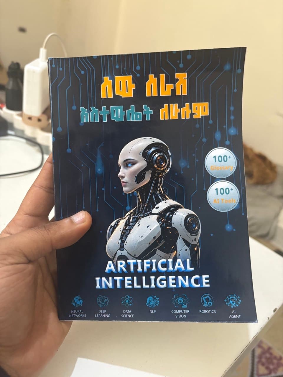 A hand holding the printed copy of አርቴፊሺያል ኢንተሊጀንስ ለሁሉም — AI FOR EVERYONE