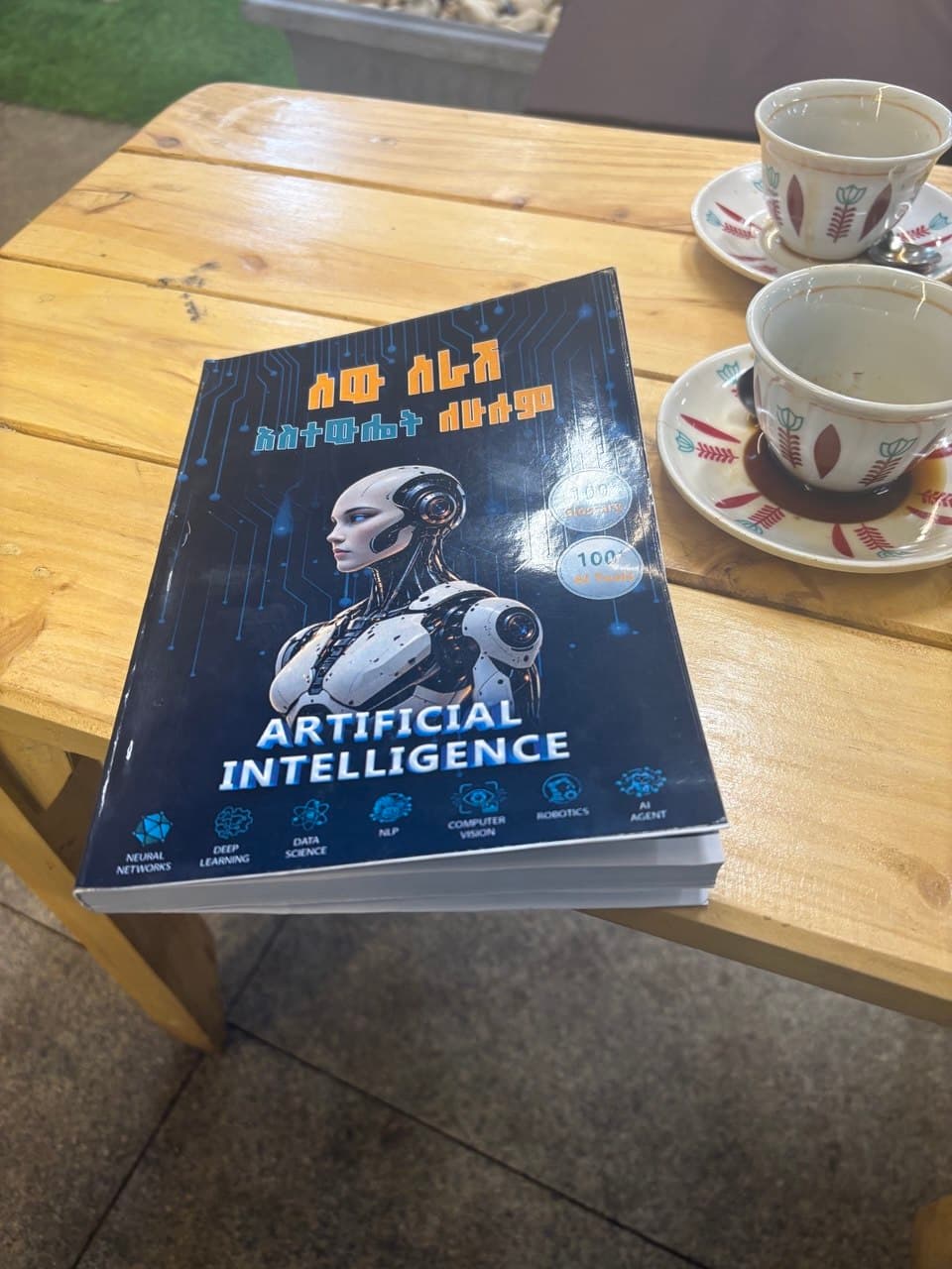 The printed AI FOR EVERYONE book on a wooden table beside traditional Ethiopian coffee cups